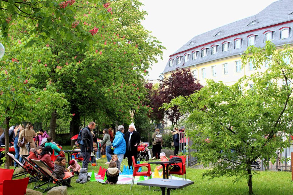 Foto vom Lesegarten mit Erwachsenen im Gespräch und in Liegestühlen sowie Kindern, die auf einer Decke auf dem Rasen spielen.