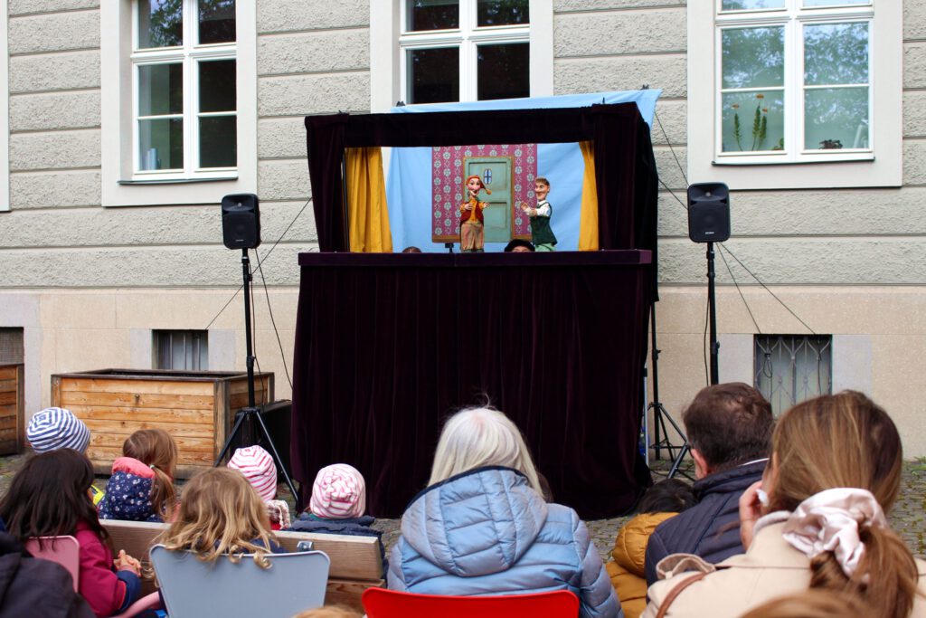 Foto der Bühne des Puppentheaters SternKundt mit zwei Kasperlefiguren und Kindern, die zuschauen.